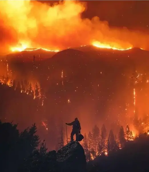 A lone firefighter silhouetted against a raging wildfire, standing on a rocky outcrop as flames engulf a forested mountainside.