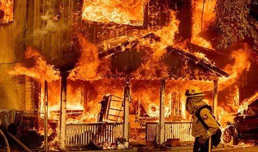 A firefighter standing in front of a house fully engulfed in flames, highlighting the devastation caused by wildfires.